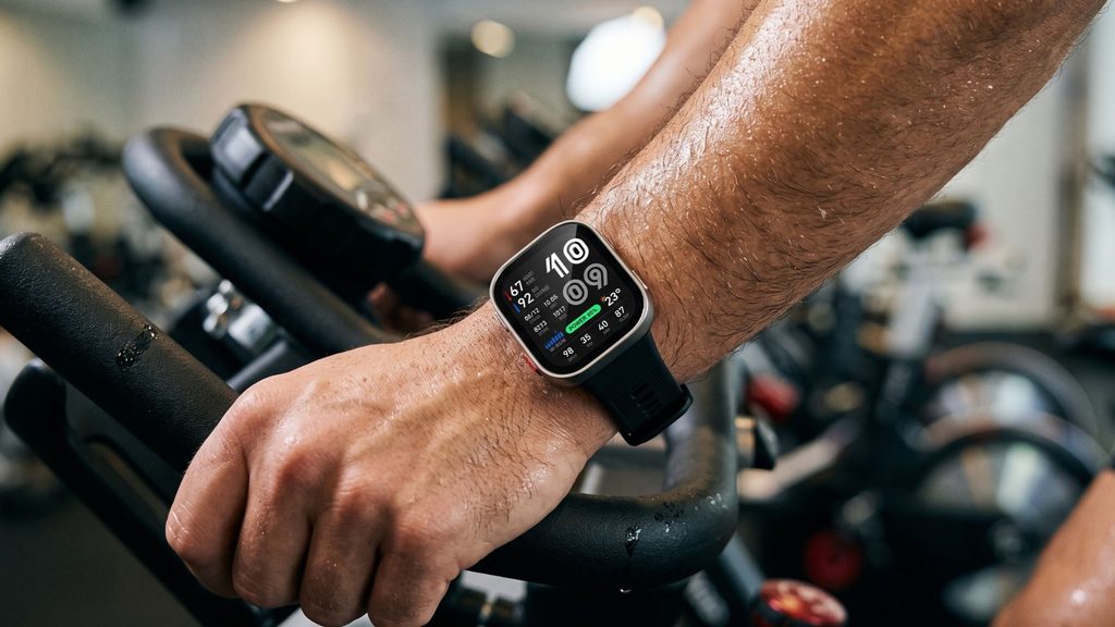 Smartwatch Amazfit Bip Max no pulso de atleta segurando o guidão de uma bike de spinning